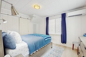 Un dormitorio con cama, lámpara de noche, ventana con cortinas, equipo de aire acondicionado y un pequeño banco de madera.