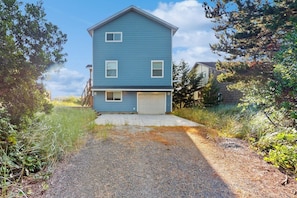 A two-story residential building with a garage, surrounded by vegetation and a gravel driveway. - 2BR oceanview with spacious deck and wifi (Copalis Beach)