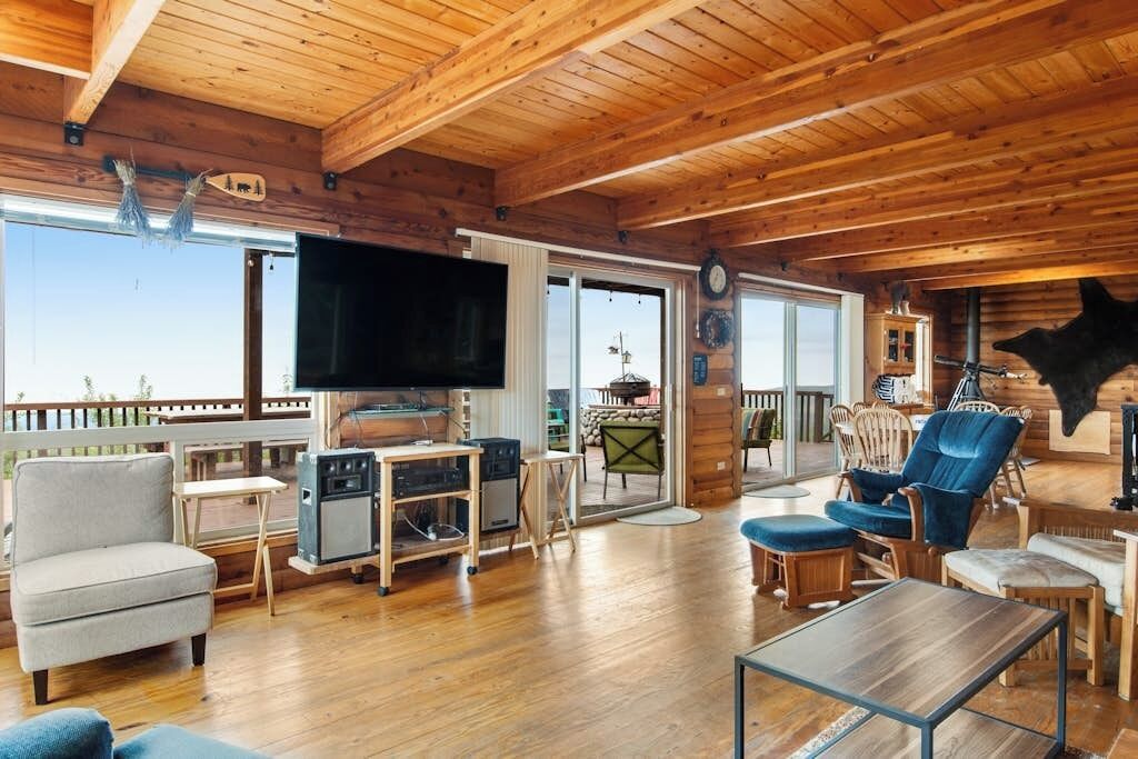 A wooden cabin interior with a TV, seating, and a large window overlooking a deck.