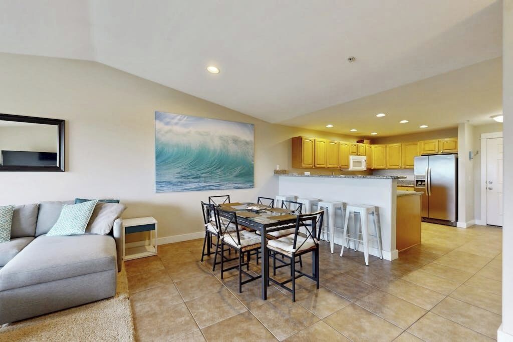 A modern living room with a gray sofa, a dining table with chairs, and a kitchen area with yellow cabinets and a stainless steel refrigerator.
