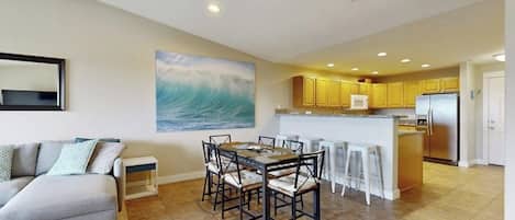 A modern living room with a gray sofa, a dining table with chairs, and a kitchen area with yellow cabinets and a stainless steel refrigerator.