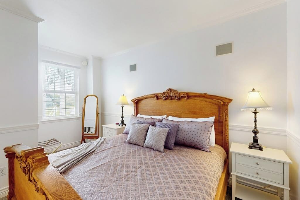 Quarto bem arrumado com cama de madeira, duas mesas de cabeceira com abajures, um espelho e uma janela com persianas.