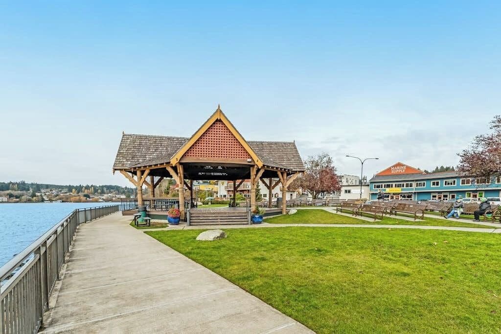 Un parc au bord de l’eau, avec un kiosque en bois, une promenade en béton et un bâtiment doté d’un toit rouge.