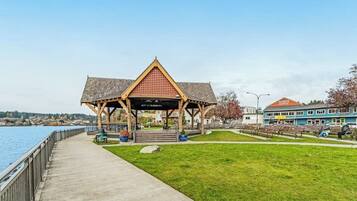 A waterfront park with a wooden gazebo, a concrete walkway, and a building with a red roof.