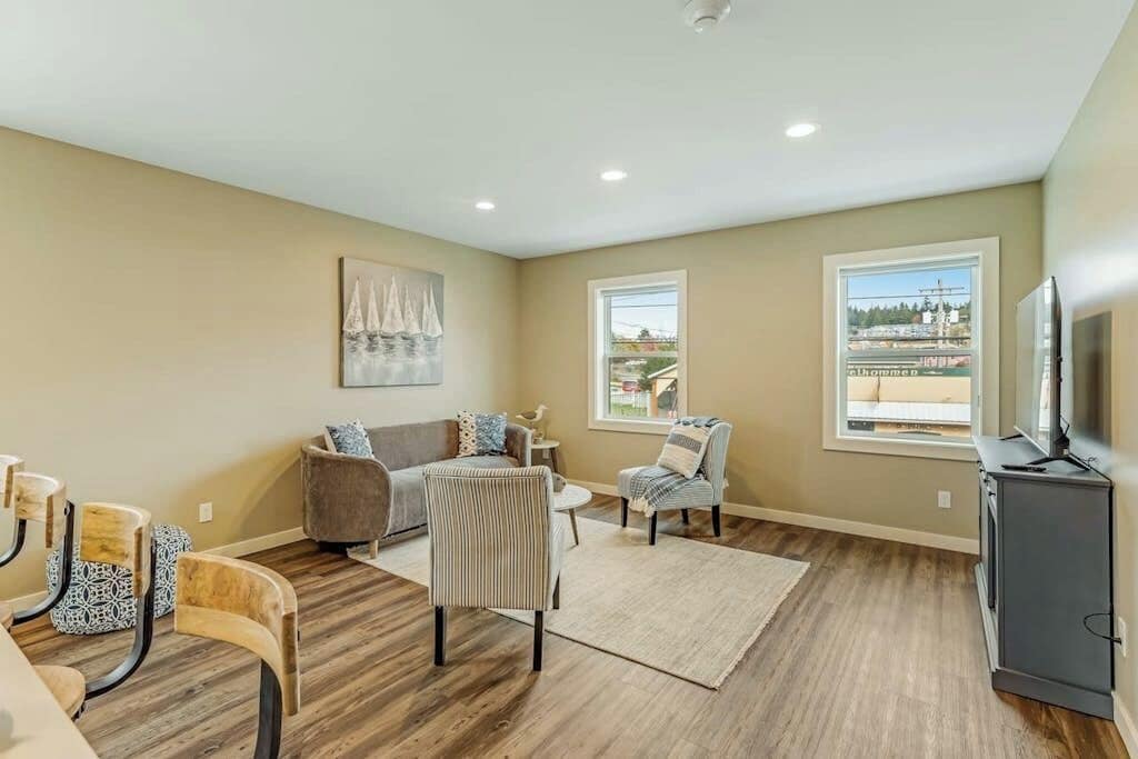 A spacious living room with a sofa, chairs, a television, and a view through the windows.