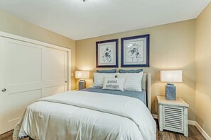 Un dormitorio con una cama, dos mesitas de noche con lámparas, una puerta y dos cuadros enmarcados con motivos florales en la pared.