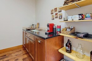 Una cuina amb armaris de fusta, una taulella, una cafetera i prestatges amb tasses i utensilis de cuina.