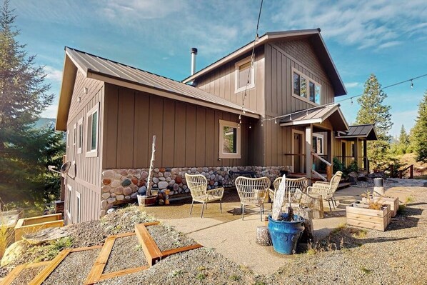 A two-story house with a stone foundation, outdoor seating area, and a clear sky. - 4BR dog-friendly retreat with hot tub & deck views (Ronald)
