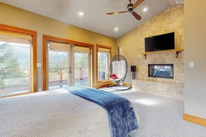 A bedroom with a large bed, a ceiling fan, a stone fireplace, and a television mounted on the wall. - 4BR dog-friendly retreat with hot tub & deck views (Ronald)