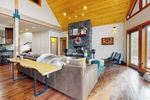 A spacious living room with a stone fireplace, wooden ceiling, and a large window. - 4BR dog-friendly retreat with hot tub & deck views (Ronald)