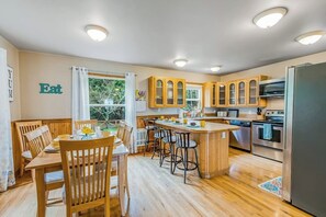 A modern kitchen with wooden cabinets, a central island, and a dining area with chairs. - Stunning waterfront 3BR with firepit & woodstove (Camano Island)