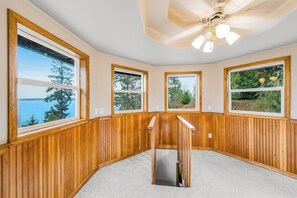 A room with wooden paneled walls, large windows with views of trees and a body of water, and a ceiling fan. - Stunning waterfront 3BR with firepit & woodstove (Camano Island)