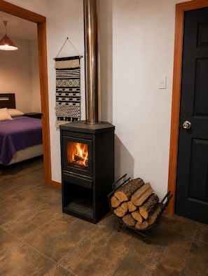 A room with a wood-burning stove, a bed with a purple blanket, and a wall-mounted tapestry. - Refugio frente al Volcán Llaima, sector captren, curacautin. (Curacautin)