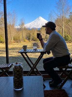 A person seated at a table with a view of a snow-capped mountain. - Refugio frente al Volcán Llaima, sector captren, curacautin. (Curacautin)