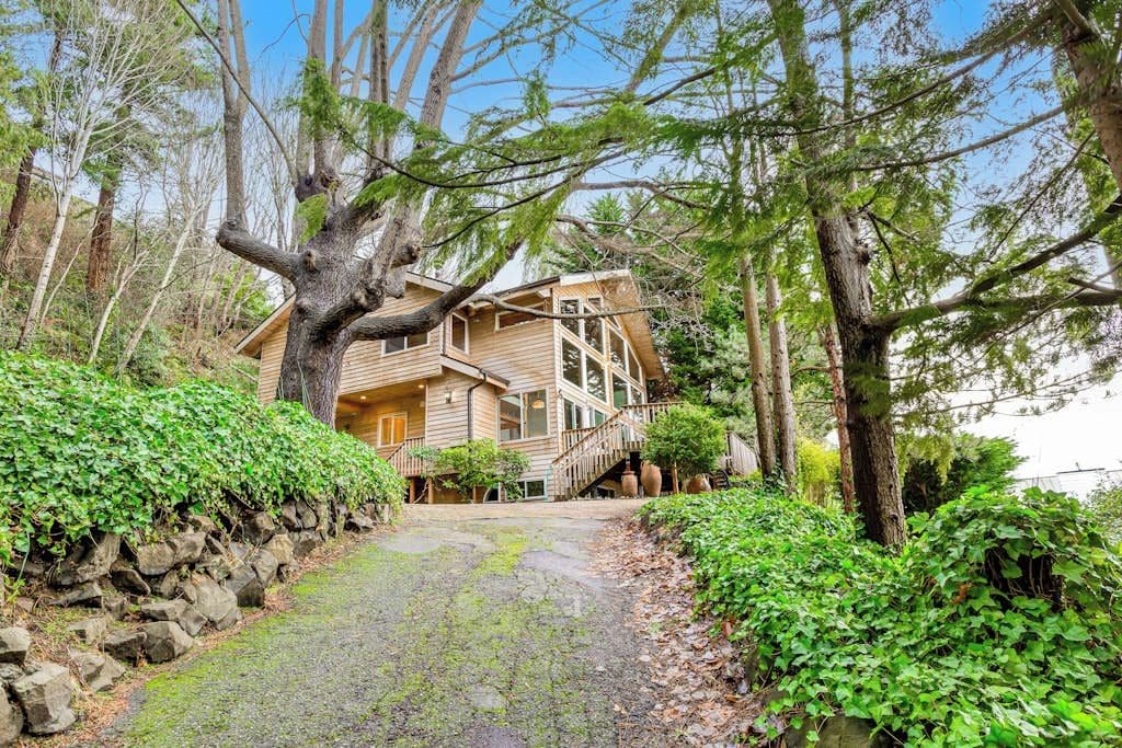 A wooden house with a staircase leading to the entrance, surrounded by trees and a stone wall.