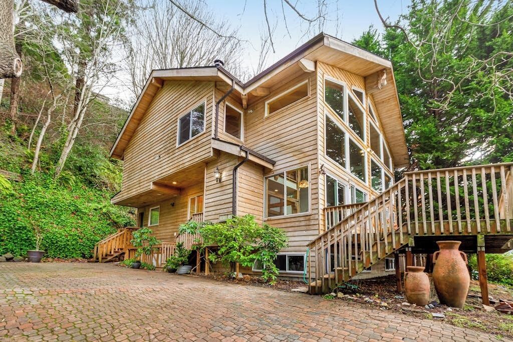 A wooden house with a staircase leading to a deck, surrounded by trees and potted plants.
