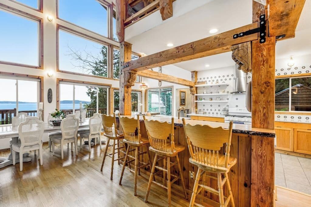 A spacious kitchen with wooden beams, a dining area with white chairs, and large windows offering a view.