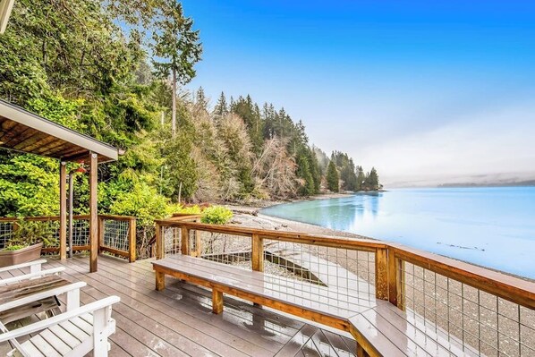 A wooden deck with a bench, overlooking a calm body of water and dense forest. - Stunning 3BR Oceanfront Dog Friendly - Dock (Tahuya)