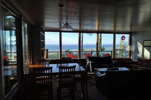 Ein Zimmer mit Esstisch, Stühlen, einer Couch und Meerblick durch große Fenster.