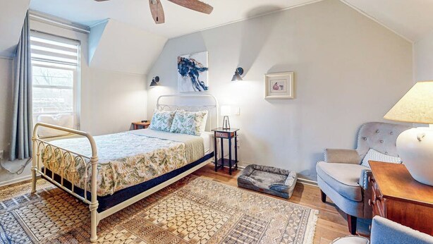 Un dormitorio con cama, ventilador de techo, mesita de noche, lámpara, silla y alfombra.