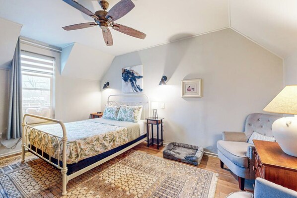 Un dormitorio con cama, ventilador de techo, mesita de noche, lámpara, silla y alfombra.