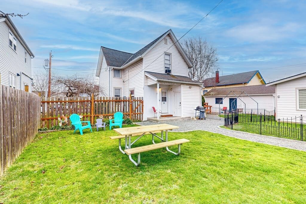 Un jardin à l’arrière d’une maison, aménagé avec une table de pique-nique et des chaises, une clôture en bois et une maison avec un garage.