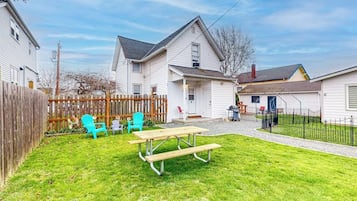 Un jardin à l’arrière d’une maison, aménagé avec une table de pique-nique et des chaises, une clôture en bois et une maison avec un garage.