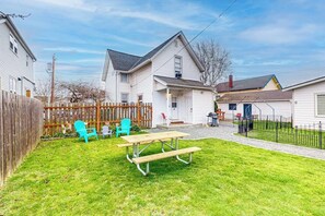 Un jardin à l’arrière d’une maison, aménagé avec une table de pique-nique et des chaises, une clôture en bois et une maison avec un garage.