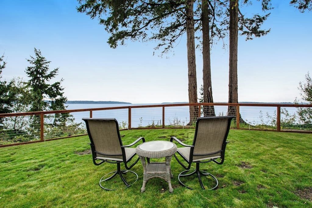 An outdoor seating area with a table and chairs, overlooking a body of water and trees.