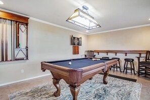 A pool table with a hanging light fixture above it, a dartboard on the wall, and a bar area with stools.