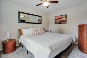 A bedroom with a bed, a round wooden nightstand, a ceiling fan, a mirror, and a painting on the wall.