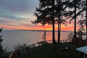 A sunset view from a deck overlooking a body of water, with trees and a small outdoor seating area.