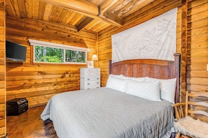 A wooden cabin bedroom with a bed, a dresser, a television, and a window with a view of trees. - Dog-friendly 2BR lakefront cabin with amazing view (Tahuya)