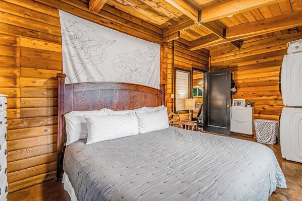 Une chambre dans une cabane en bois, avec un lit, une grande tapisserie à motifs floraux, une commode et un réfrigérateur.
