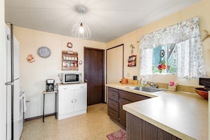 A compact kitchen with a microwave, coffee maker, and a small refrigerator. There is a window with a floral curtain and a view of greenery outside. - Large 2BR Oceanfront - Deck - Firepit - Washer Dryer (Camano Island)
