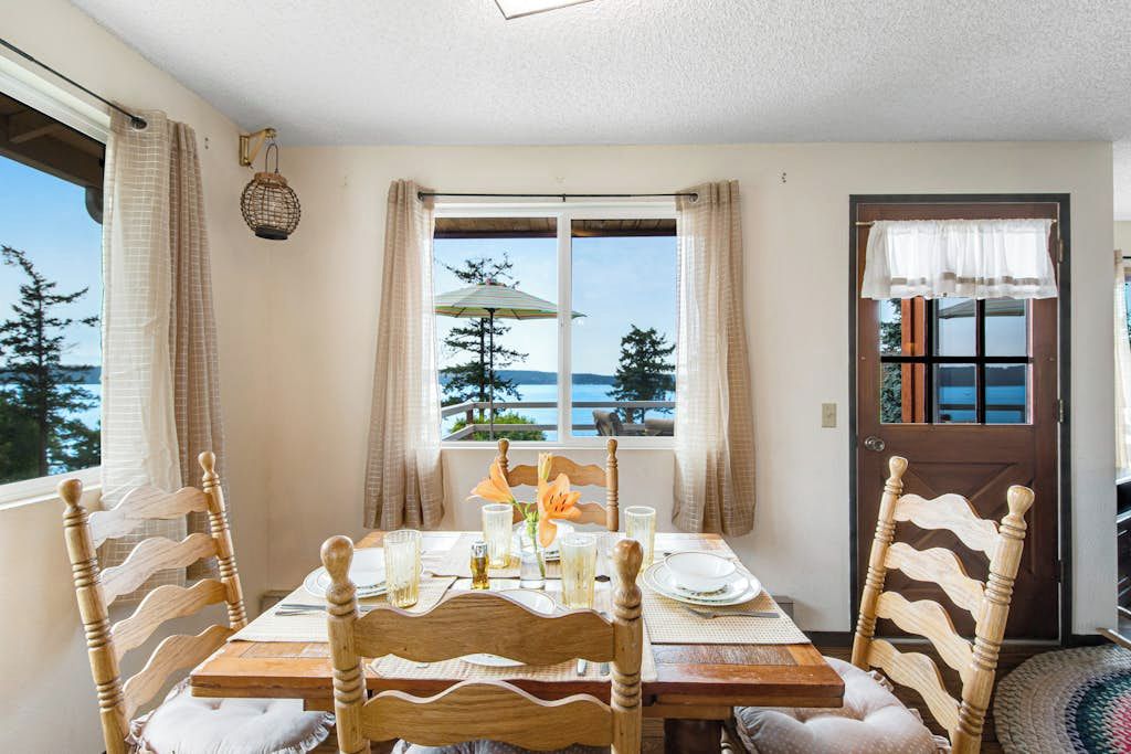 A dining area with a table set for four, wooden chairs, a view of the sea through a window, and a door with a curtain.
