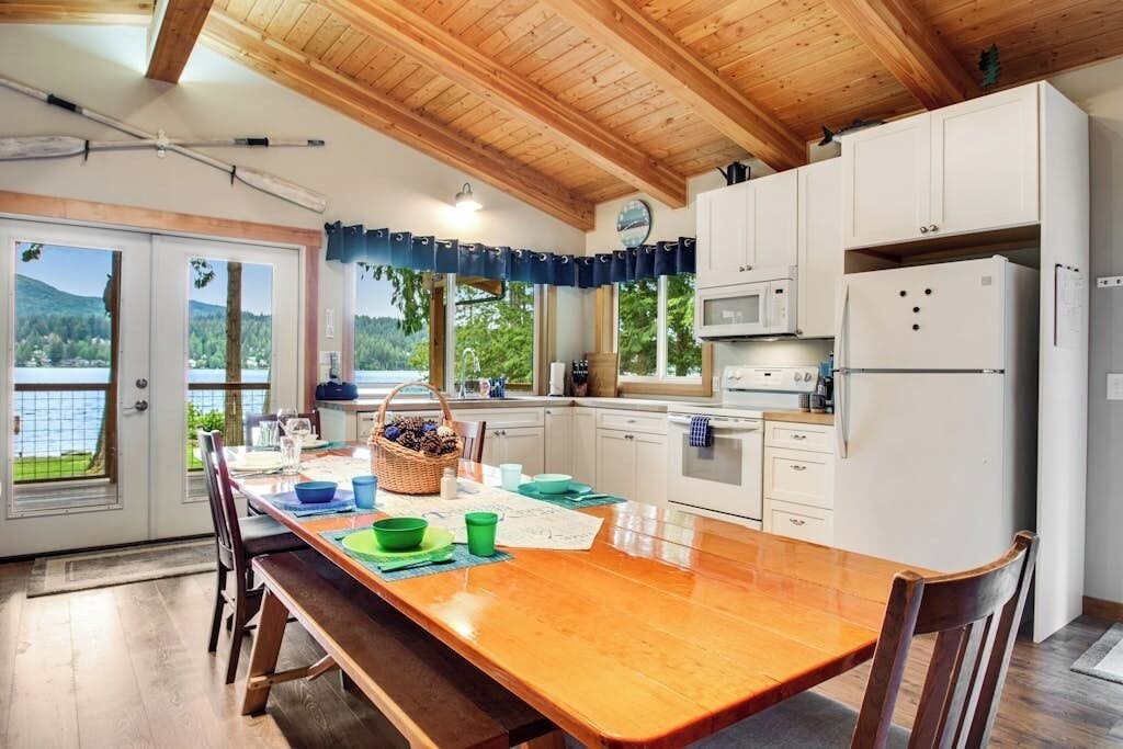 Une cuisine avec une table en bois, des placards blancs et une vue sur l’extérieur.