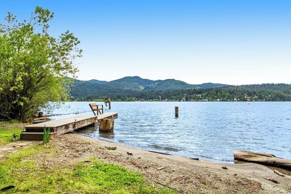 Une jetée au bord du lac, avec un banc et une table, entourée de verdure et avec des montagnes en arrière-plan.