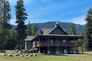 A two-story lodge with a balcony, surrounded by a stone wall and trees, set against a mountainous backdrop. - 4BR Home with fire pit & pickleball and basketball court (Cle Elum)