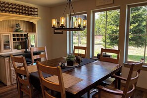 A dining area with a wooden table and chairs, a chandelier, and a view of the outdoors through large windows. - 4BR Home with fire pit & pickleball and basketball court (Cle Elum)