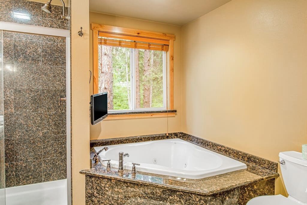 A bathroom with a large bathtub, a toilet, and a window with a view of trees.