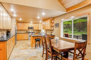 A kitchen with wooden cabinets and a stone floor, a dining table with chairs, and a large window with a view of greenery. - 2BR cabin with patio & grill - close to the lake (Ronald)