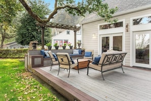 An outdoor deck with seating and a pergola, surrounded by a garden and a house.