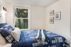 A bedroom with a large window, a bed with blue bedding, and three framed pictures on the wall.