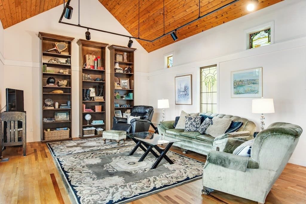 A spacious living room with a high wooden ceiling, a green sofa, a black leather chair, and a patterned rug.