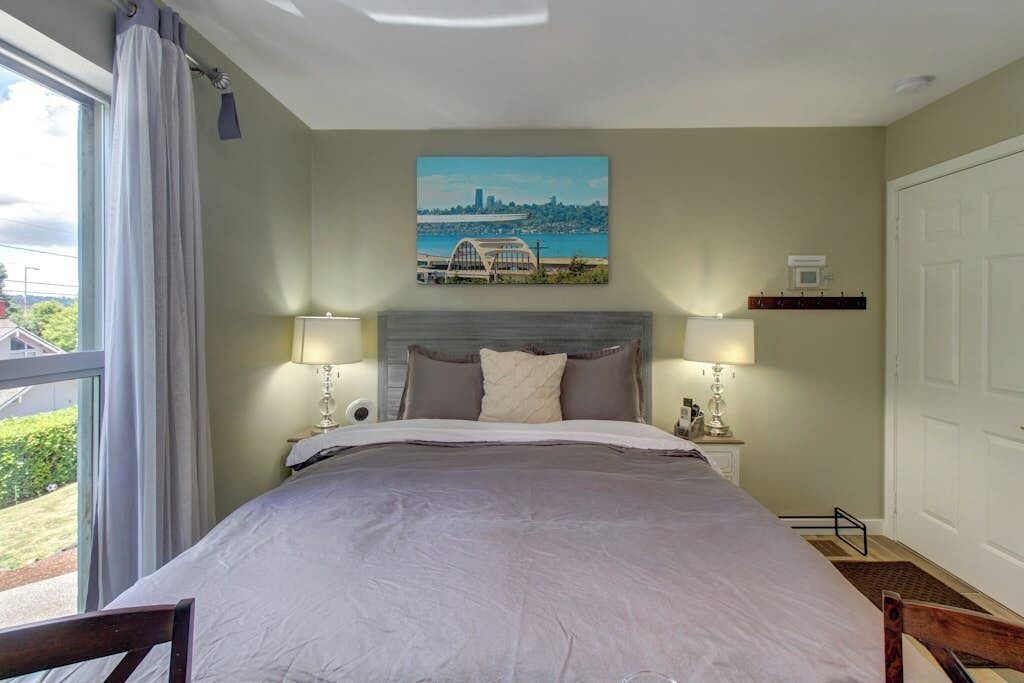 A hotel room with a large bed, two bedside lamps, a painting on the wall, and a view of the outdoors.