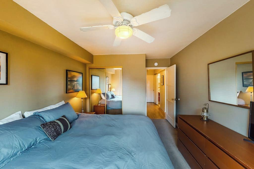 A bedroom with a bed, bedside lamps, a dresser, a ceiling fan, and a hallway leading to another room.