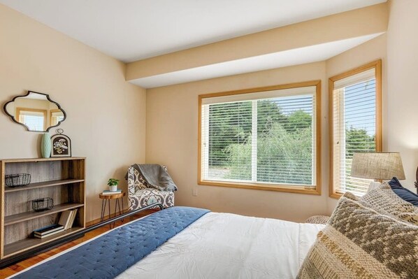 A bedroom with a large window, a bed with a blue and white duvet, a side table with a lamp, and a bookshelf.