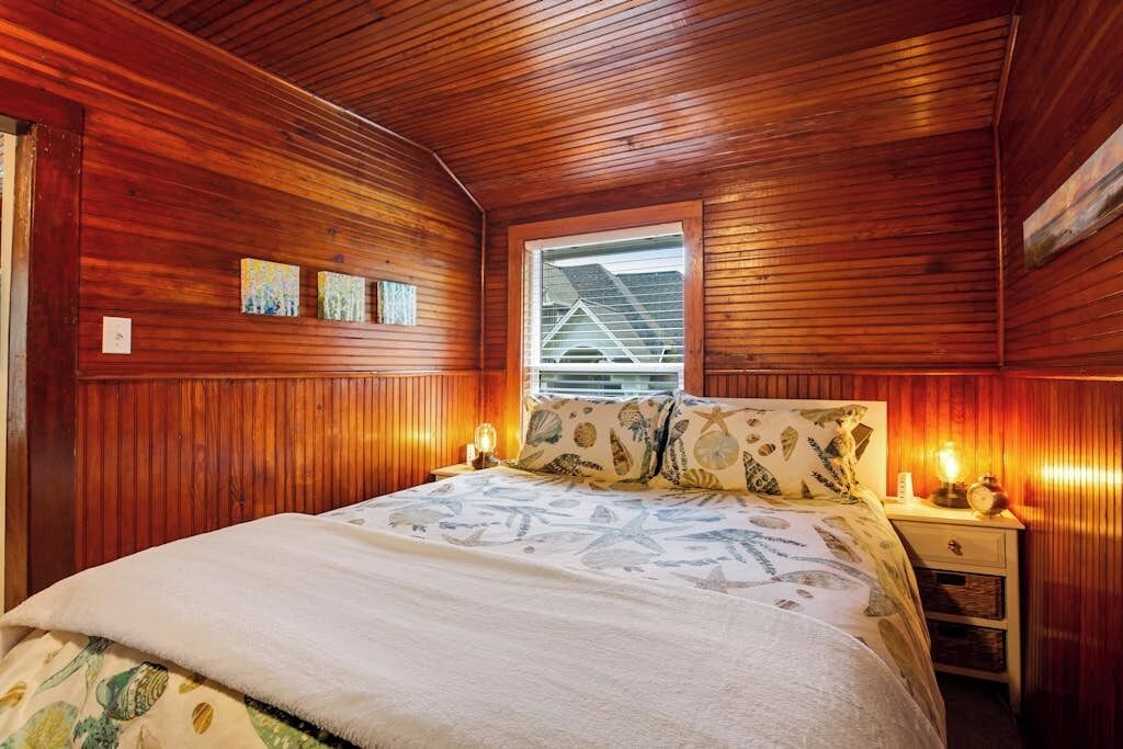 A bedroom with wooden paneling, a bed with a patterned bedspread, two bedside lamps, and a window with blinds.