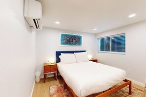 Ein Schlafzimmer mit einem Bett, Nachttischen, einer Wand-Klimaanlage, einem Fenster mit Jalousien und einer Deckenleuchte.